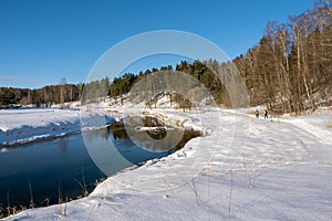 Landscape with a small river and two skiers on a winter sunny day and blue sky