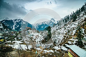 A landscape of a snow capped valley in a mountain with pine trees and houses. Blue sky with clouds above