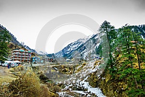 A landscape of a snow capped valley with trees and houses. Mountain with snow capped peaks and blue sky in the background