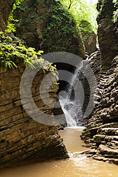 Landscape with summer stormy cascade waterfalls in canyon, lush green forest, sunbeams, splashes, layered rocks overgrow green.