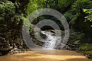 Landscape with summer waterfall in lush green forest with sunbeams, splashes and layered rocks overgrow green moss on mountain.