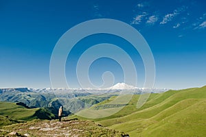 Landscape with a view of Elbrus and green meadows. A man looks alone at the snowy mountains