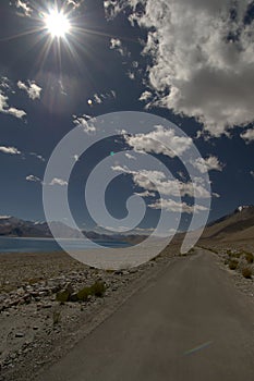A landscape view on the way to Pangong Tso lake with empty road under cloudy-blue skies and sunburst, Ladakh, India.