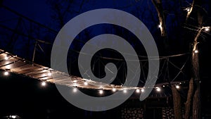 The landscape of a wooden bridge lit in the darkness of the light bulb night, surrounded by the silhouettes of the trees