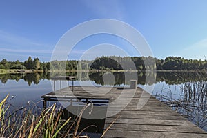 Landscape with wooden long jetty with chair for fishing, lake, forest on horizon and clear blue sky in summer