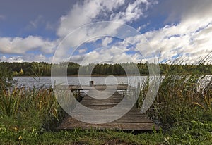 Landscape with wooden long jetty with chair for fishing, lake, forest on horizon and cloudy blue sky in summer