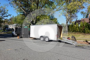 A landscaping truck with a long white enclosed trailer trailer with it`s rear ramp down seen on a shady residential asphalt stree