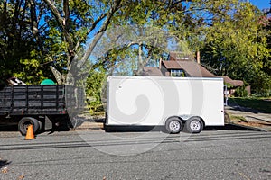 A landscaping truck with a long white enclosed trailer trailer seen on a shady residential asphalt street