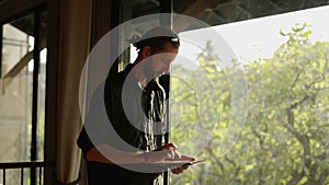 A lanky bearded man looking at the window with his tablet in his hand