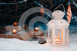 Lantern with a burning candle and wrapped gifts in the snow under the Christmas tree on Christmas Eve