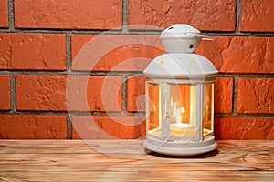 A lantern with a glowing candle in on the wooden table, brick wall background