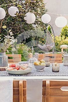 Lanterns above table with food and drink in the garden with plants and wooden chairs. Real photo