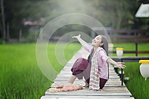 Lao beautiful young woman wearing local dress sitting on wooden bridge in green rice field happily.