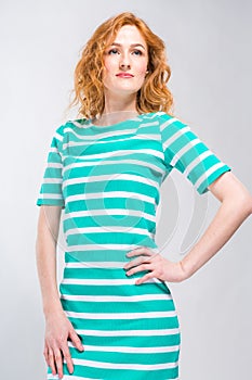 A lap portrait of a young, beautiful woman with red curly hair in a summer dress with stripes of blue in the studio on a gray back