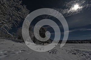 Lappish moonlit winter landscape with forest and lake on blue sky with full cloud covered moon