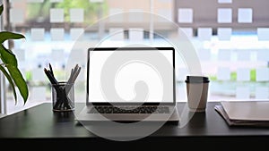 Laptop with blank screen, pencil holder, coffee cup and books on black wooden table.