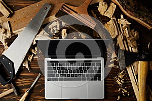 laptop with blank screen surrounded by axe, handsaw, chisel, paintbrush and hammer on wooden table