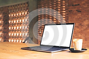 Laptop with blank white desktop screen and coffee cup on wooden table with brick wall background