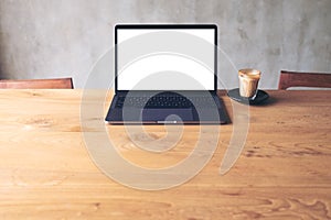 Laptop with blank white desktop screen and coffee cup on wooden table with concrete wall background