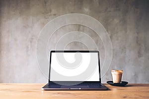 Laptop with blank white desktop screen and coffee cup on wooden table with concrete wall background