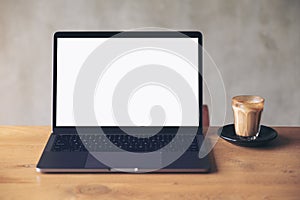 Laptop with blank white desktop screen and coffee cup on wooden table with concrete wall background