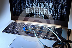 Laptop with blue matrix and message SYSTEM HACKED on the screen as cybercrime symbol, stethoscope on the keyboard, concept for