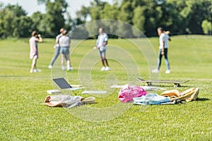 laptop books backpacks and skateboard on grass and teenagers playing behind