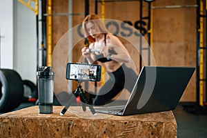 Laptop, bottle and smartphone on tripod is on the table. Beautiful strong woman is in the gym