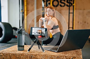 Laptop, bottle and smartphone on tripod is on the table. Beautiful strong woman is in the gym