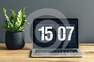 Laptop with clock screensaver placed on wooden desk with fresh green potted plant