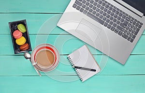 Laptop, colored macaroons cookies, cup of coffee, notebook with a pen on a blue wood table. Top view. Flat lay.