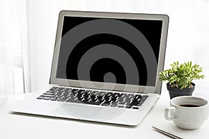 Laptop computer with black blank mockup screen on top of white office desk table with coffee cup and pen