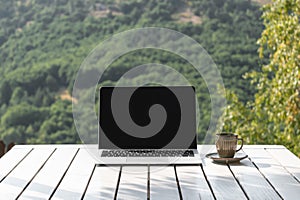 Laptop computer with black screen and coffee cup on table in outdoor cafe of mountain resort. Online work and relax