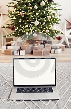Laptop computer with blank white mockup screen over Christmas tree and gift boxes at background