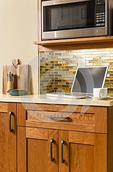 Laptop computer and coffee cup on counter in contemporary home kitchen with wood cabinets and glass tile backsplash