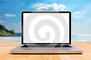 Laptop computer with empty blank white screen for copy space on wooden desk with blurry summer sea ocean beach at background.