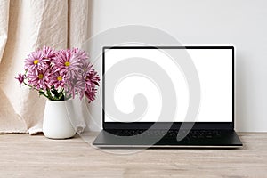 Laptop computer empty screen mockup, vase with pink daisy flowers bouquet on beige wooden table, white wall and linen