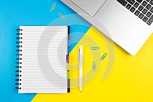 Laptop computer, open paper notebook and white pen on blue and yellow color background. Top view, copy space for text