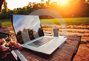 Laptop computer on rough wooden table with coffee cup and bouquet of peonies flowers in outdoor park