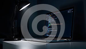Laptop displaying programming code illuminated by desk lamp in dark room