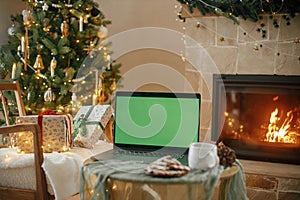 Laptop with empty screen and stylish christmas gifts on background of fireplace, tree with golden lights in festive cozy room.