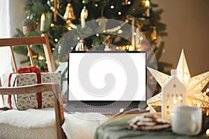 Laptop with empty screen and stylish christmas gifts on modern chair in festive decorated room with tree, golden lights and star.