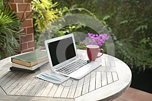 Laptop, face mask, books and red cup on top of table next to garden