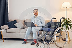 With laptop on the legs. Disabled man in wheelchair is at home