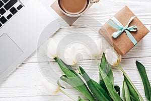 laptop with morning coffee and tulips and stylish gift box on white wooden rustic background. flat lay freelance, work gadgets wi