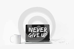 laptop with Never give up inspiration on screen, earphones and white mug