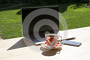Laptop, phone and beautiful porcelain cup of tea stand on light wooden table in green garden.