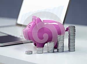 Laptop and a pink piggy bank, ceramic shiny, on wooden white desk, with stacks  of 50 cents USA coins, 3d render