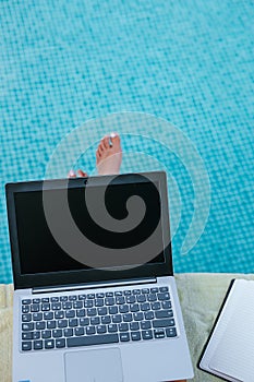 laptop propped up on one\'s legs at the edge of the pool, accompanied by a notebook for notes
