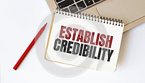 Laptop, red pen and notepad with text ESTABLISH CREDIBILITY in the white background photo